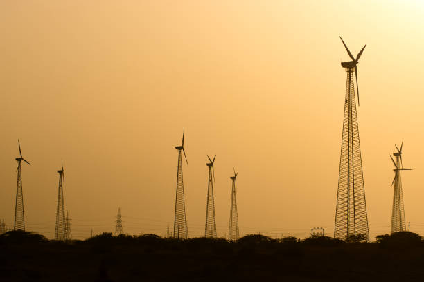 This image captures a striking scene of wind turbines set against the backdrop of a vibrant sunset. The silhouettes of the tall, sleek turbines are sharply defined against the warm, golden-orange sky, where the sun is positioned just above the horizon. The descending sun casts a soft, diffused light across the landscape, enhancing the serene and almost ethereal atmosphere of the image.The turbines, with their slender, lattice-style towers, stand tall and majestic, symbolizing the modern pursuit of renewable energy. The simplicity of the composition, with the focus on the turbines and the setting sun, emphasizes the harmony between technological advancement and the natural world.The scene likely depicts a wind farm in a remote, open area, where the expansive sky meets the low horizon dotted with sparse vegetation. The image beautifully illustrates the convergence of nature and technology, highlighting the role of sustainable energy solutions in preserving the beauty of our environment.