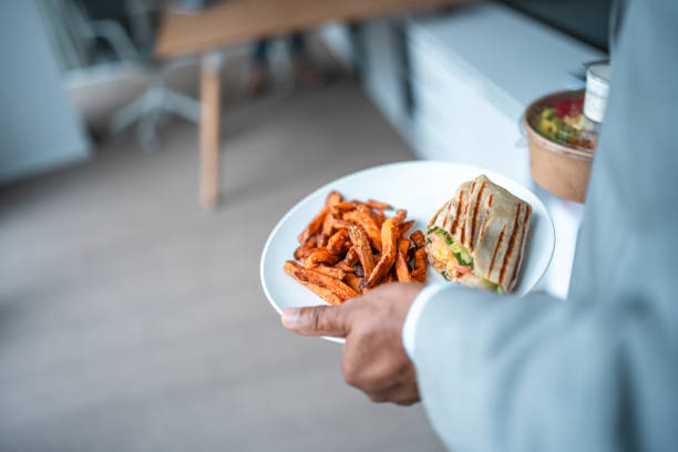 An elderly Hispanic businessman in professional attire holds a plate with a sandwich and sweet potato fries, approaching his office desk to enjoy a meal.