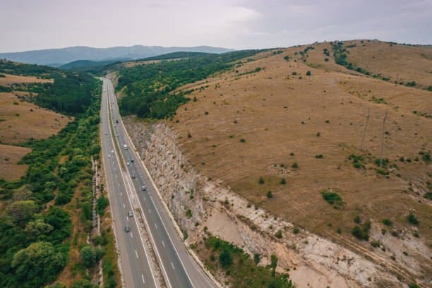 Aerial view of highway traffic between green trees and a cliff with a plateau of dry grass on a cloudy summer day.