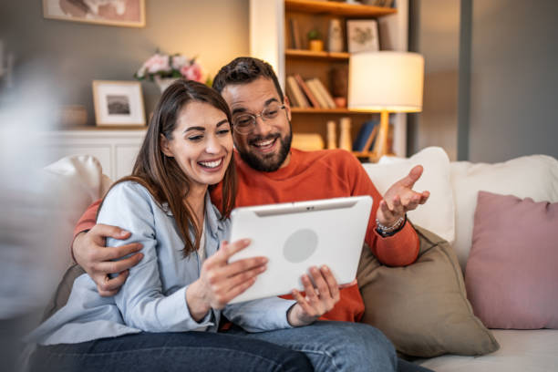 istock A couple laughs while using a digital tablet together at home. Boy and girl relaxing and using social media on digital tablet in living room at home 2166907972