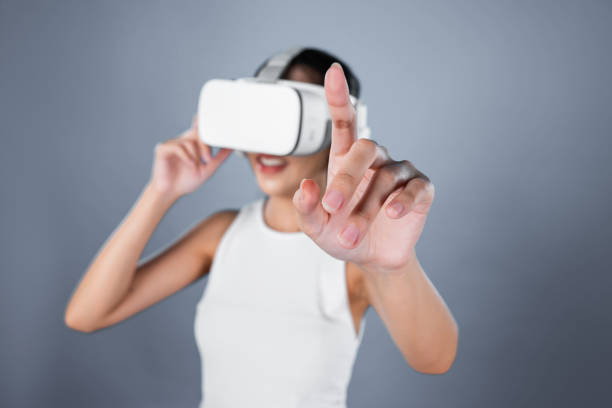 Smart female standing with gray background wearing VR headset connecting metaverse, futuristic cyberspace community technology. Elegant woman excited seeing generated virtual scenery. Hallucination.
