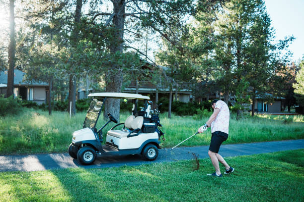 An active Caucasian man swings his club while playing golf on the course in front of a golf cart on a beautiful, sunny day.