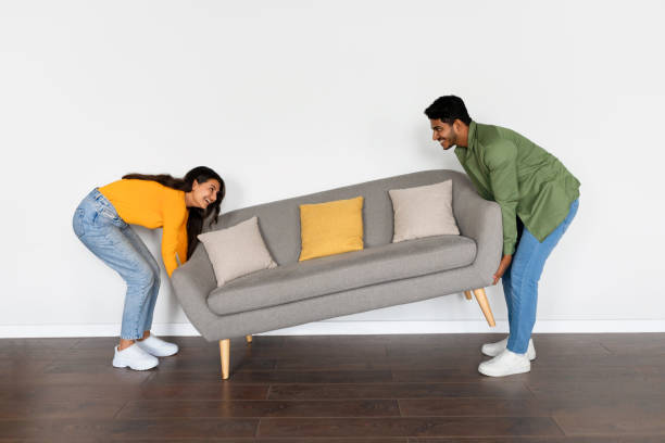 istock Indian spouses moving grey sofa, white wall background 2166926156