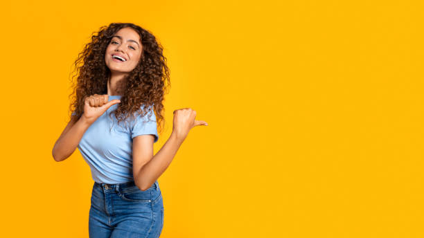 istock Joyful woman pointing aside with thumb on yellow 2166926365