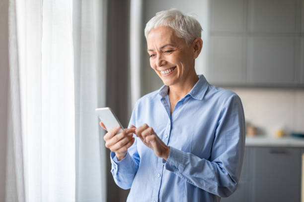 istock Smiling Senior Lady Using Mobile Phone Texting Standing At Home 2166927902