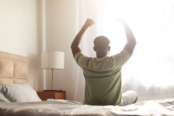 istock black guy sitting on bed indoor stretching body and arms 2166928118