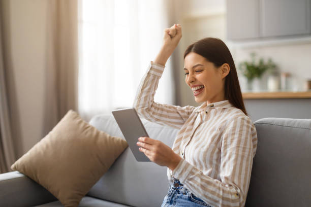 istock Joyful young woman celebrating success with tablet on couch 2166929593
