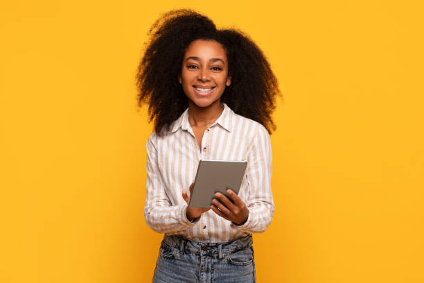 istock Smiling black woman holding tablet on bright yellow background 2166932125