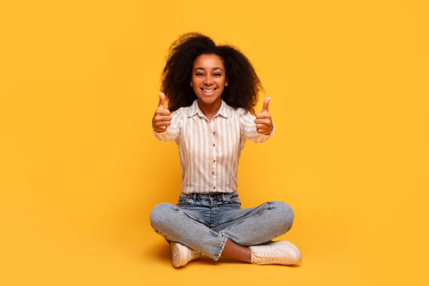 istock Happy black woman giving thumbs up while sitting on yellow backdrop 2166932127