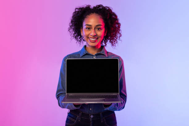 istock Smiling black lady presenting laptop with blank screen, denim attire, neon background 2166933514