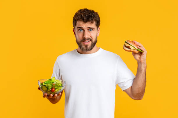 istock Confused Guy Holding Burger And Vegetable Salad Choosing Diet, Studio 2166933792