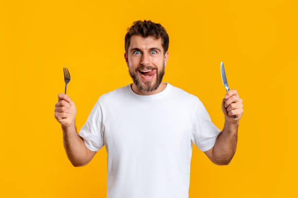Hungry young guy holding knife and fork on yellow background