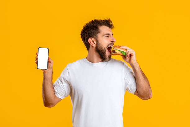 istock Hungry man with burger shows smartphone blank screen, yellow background 2166933794