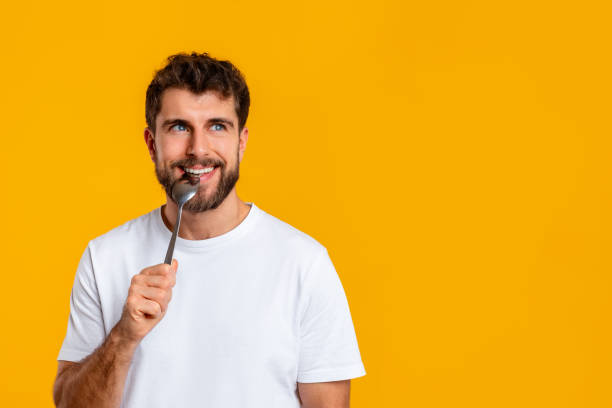 Hungry guy holding and biting spoon looking at free space aside, posing on yellow studio background. Portrait of funny young man advertising food offer, waiting for dinner. Male nutrition concept