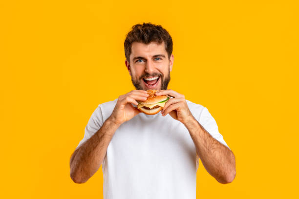 istock man biting delicious takeaway cheeseburger smiling to camera, yellow backdrop 2166933842