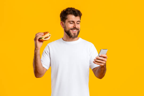 istock Cheerful young man holding burger using cellphone over yellow background 2166933849