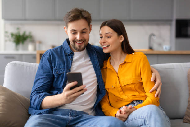 istock Spouses smiling and looking at smartphone together, sitting on sofa 2166934764