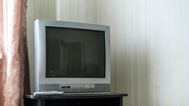 istock Old-fashioned CRT television set sits on dark table in room with patterned wallpaper and partially drawn curtains. Remote control is placed beside TV adding nostalgic touch to simple interior 2166938020