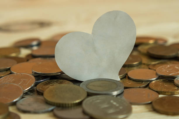 Coins from different countries are scattered on the wooden table. On their background is a white heart. Selective focus. The concept of love money