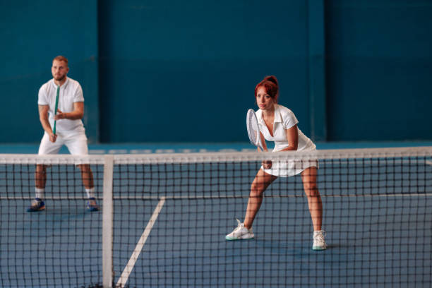 Scene from a mixed-doubles tennis match on a blue indoor tennis court with the female player close to the net and the male player behind her.