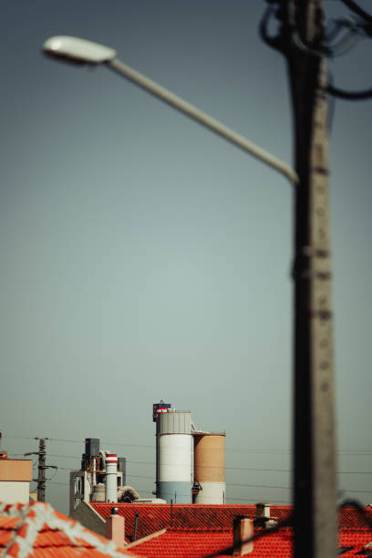 A telephoto distant view of industrial silos against a clear sky, partially obscured by an out-of-focus streetlight in the foreground, with rooftops in the lower section