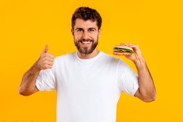 istock cheerful guy holding cheeseburger gesturing thumbs up on yellow background 2166948562