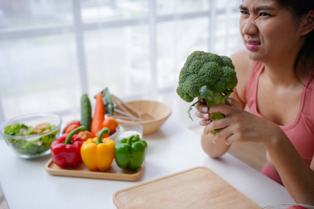 istock Young woman is making a disgusted face while reluctantly holding broccoli over a table with other healthy food 2166954489