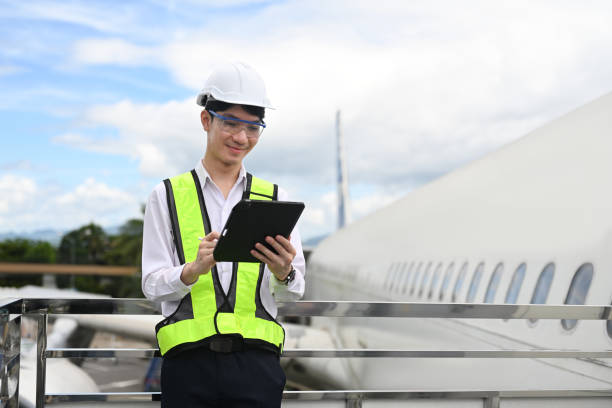 istock Young man wearing yellow vest and hard hat using digital tablet, standing on metal railing next to an airplane 2166967568