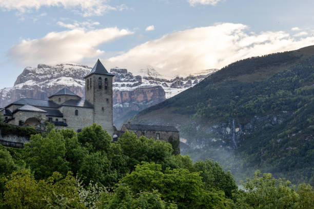 Scenic view of Torla, a charming village nestled in the Aragonese Pyrenees, Spain. The ancient stone church stands prominently, surrounded by lush greenery and set against a backdrop of snow-capped mountains under a partly cloudy sky. vista panorámica de torla, un encantador pueblo enclavado en el pirineo aragonés, españa. la antigua iglesia de piedra ocupa un lugar destacado, rodeada de exuberante vegetación y con un telón de fondo de montañas nevadas bajo un cielo parcialmente n