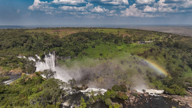 Kalandula Waterfall flows powerfully amidst vibrant vegetation with a rainbow forming in the mist on a sunny day.