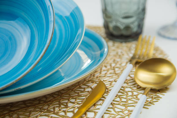 istock Bright blue serving plates with a spiral pattern, tilted one after the other, serving devices of gold - white color are located on the side, glasses and glasses are blurred behind them, the side view is large on a white background. 2166977619