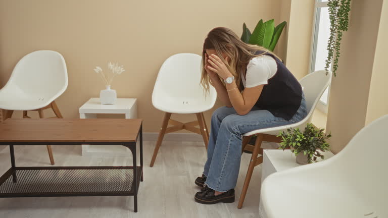 A young woman sitting in a modern indoor waiting room, appears stressed or anxious with head in hands, surrounded by minimalist white chairs and plants.
