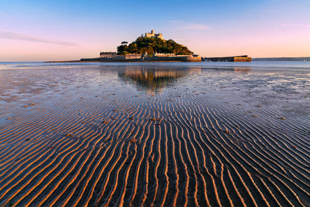 A scenic view of a castle on a hill surrounded by lush greenery, with a sandy beach in the foreground and cloudy skies above.