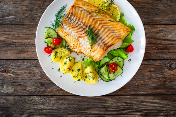 istock Fried cod fillet, boiled potatoes, fresh vegetables and lemon slices served on white plate on wooden table 2166986526
