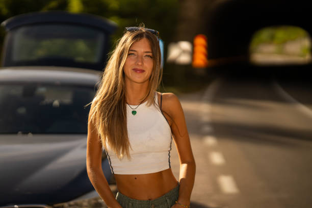 Portrait of young woman standing in front of a car on a road and looking at camera on sunny summer day