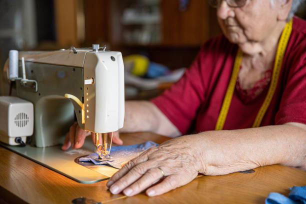 An elderly woman working diligently at a vintage sewing machine. She is focused on her task, with glasses on and a tape measure draped around her neck, sitting in a cozy, warmly lit room.