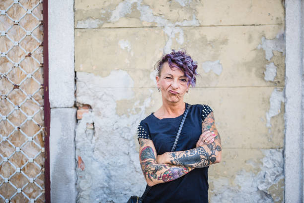 A confident woman with vibrant tattoos and purple hair stands with her arms crossed in front of a weathered wall.