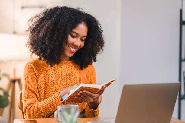 istock Smiling African American woman reading a notebook while working at home, sitting at a desk with a laptop and a coffee mug. 2167005995