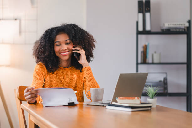 istock African American woman multitasking at desk, speaking on the phone while reviewing documents, with a laptop at home. 2167006052