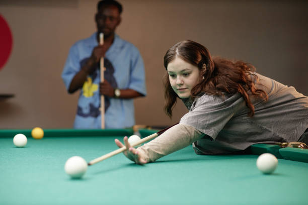 Young Caucasian woman intently aiming and playing billiards in a game room setting under indoor lighting with people in background conversing and enjoying activities