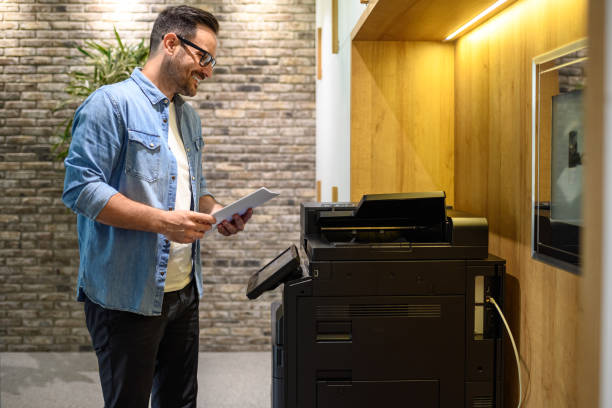 istock Smiling young businessman in glasses using computer printer for photocopying documents in modern office 2167029862