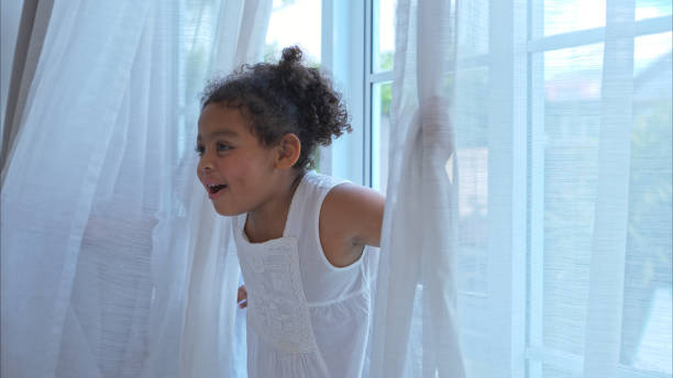 A tiny girl plays hide-and-seek with drapes in her bedroom