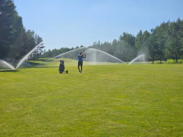 A golfer swings on a well-maintained green while sprinklers provide water on a sunny afternoon.