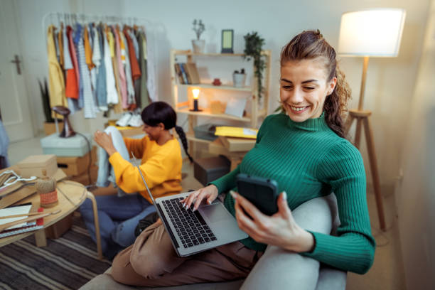 istock Young cheerful woman using phone and laptop while working with her friend at home based clothing store 2167062847