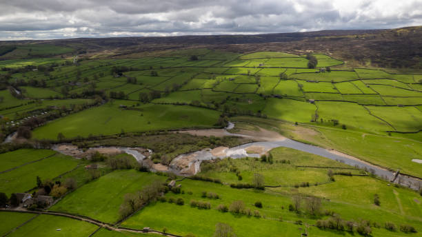 istock Aerial view of a lush green countryside with a winding river, patchwork fields, and overcast skies. 2167064221