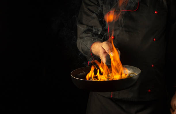 istock Chef cooking vegetables on a burning frying pan with flame. Restaurant and hotel service concept on a black background. Flambe or cooking with natural fire 2167067455