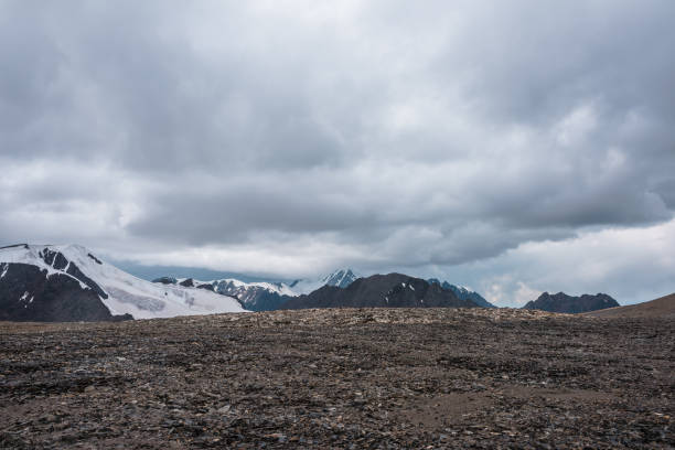 istock Dramatic misty top view from stone pass to big glacier tongue among sharp rocks and large snow-capped pinnacle in rainy low clouds. Dark atmospheric mountain silhouettes in rain in gray cloudy sky. 2167078891