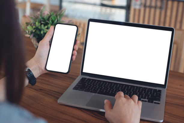 istock Mockup blank white screen cell phone and laptop for web template or mobile app design. Woman hand using mobile phone during working on laptop computer on wooden table at coffee shop or home office 2167092641
