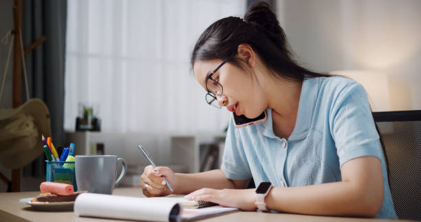 istock Young woman wearing glasses is talking on the phone while writing down work details. 2167094034