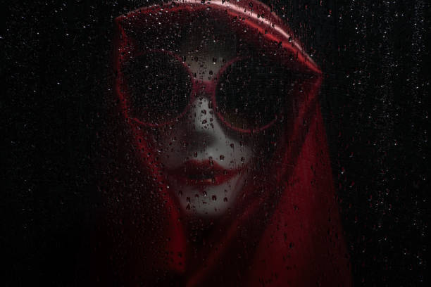Plastic model head with home painted mask wearing red silk wrap home made mask and large sunglasses behind water droplet covered window Asia Thailand